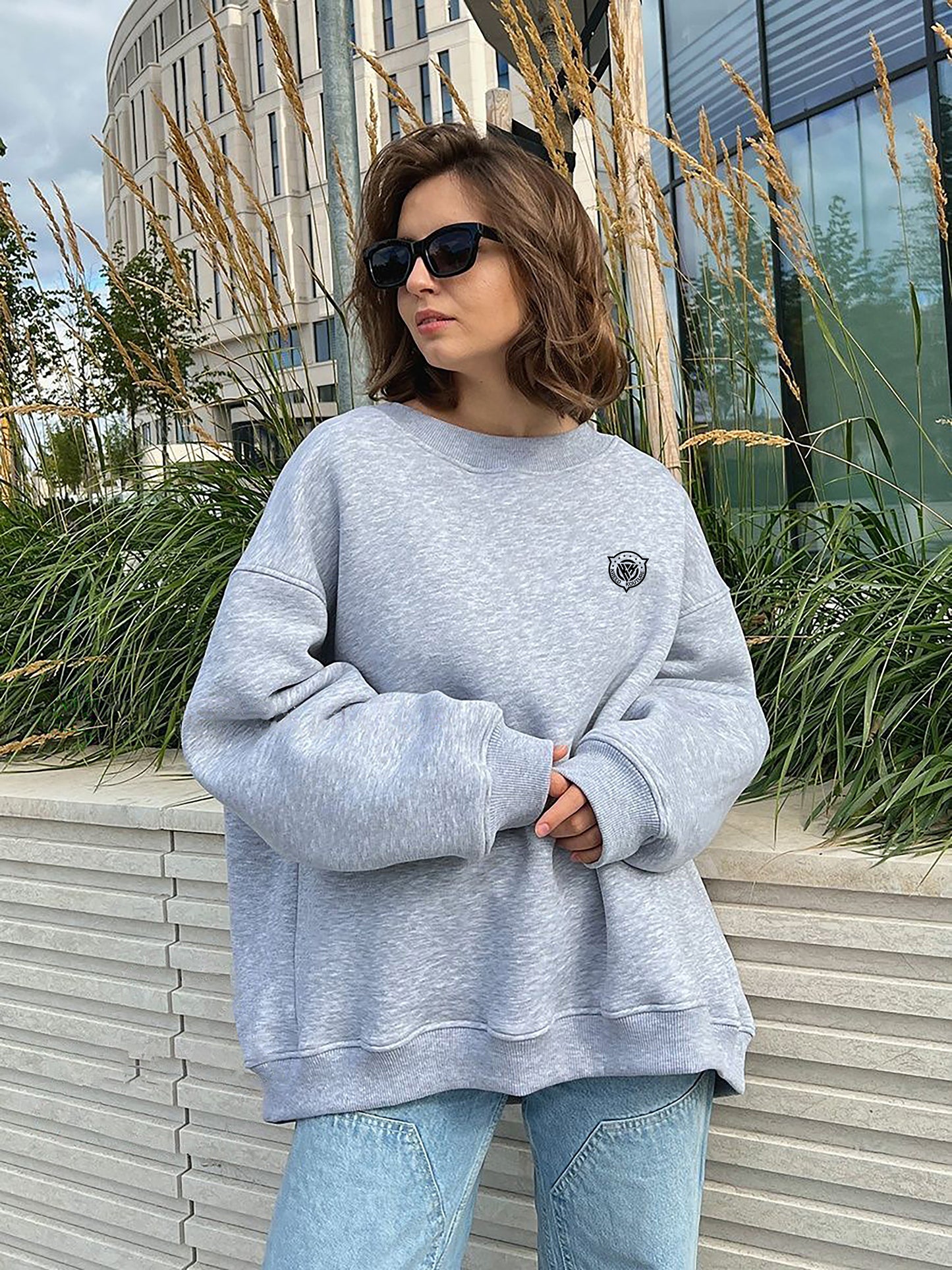 Woman wearing a gray sweatshirt and sunglasses standing outdoors with plants and a building in the background