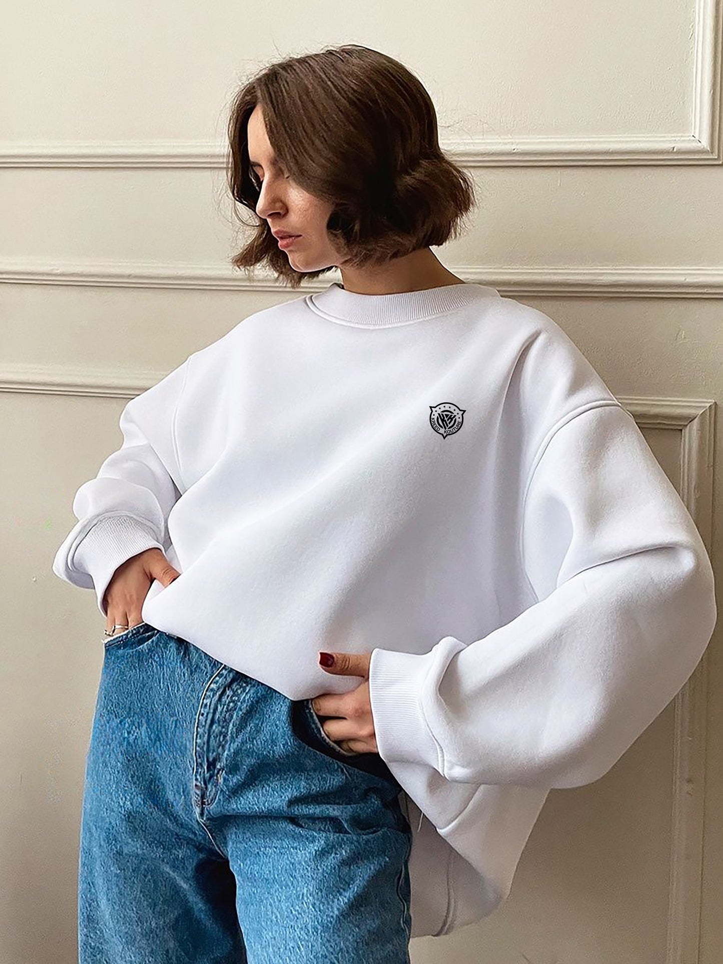 Person wearing a white sweatshirt with a small logo and blue jeans against a neutral wall.